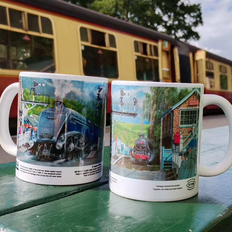 Two white ceramic mugs printed with vintage train station scenes, displayed on a green table with a blurred train and outdoor background.