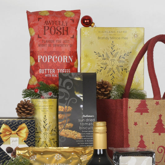 A curated gift basket or hamper arrangement featuring a red bag of Awfully Posh Popcorn, a yellow box of Highland Fayre Brandy Mince Pies, a pack of Bateman's sun dried tomato biscuits, a red and beige just bag, styled with pinecones and evergreen branches on a light gray background.