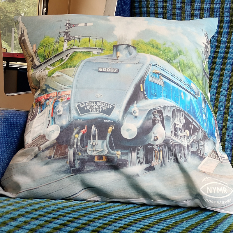 A square decorative cushion featuring a printed illustration of a blue and cream steam locomotive train, resting on a blue and green plaid fabric surface.