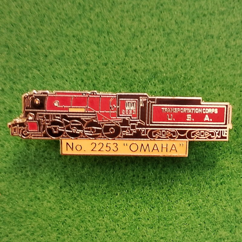 A small train-themed enamel pin or badge shaped like a steam locomotive with a plaque reading "No. 2253 'OMAHA'" on a green textured background.