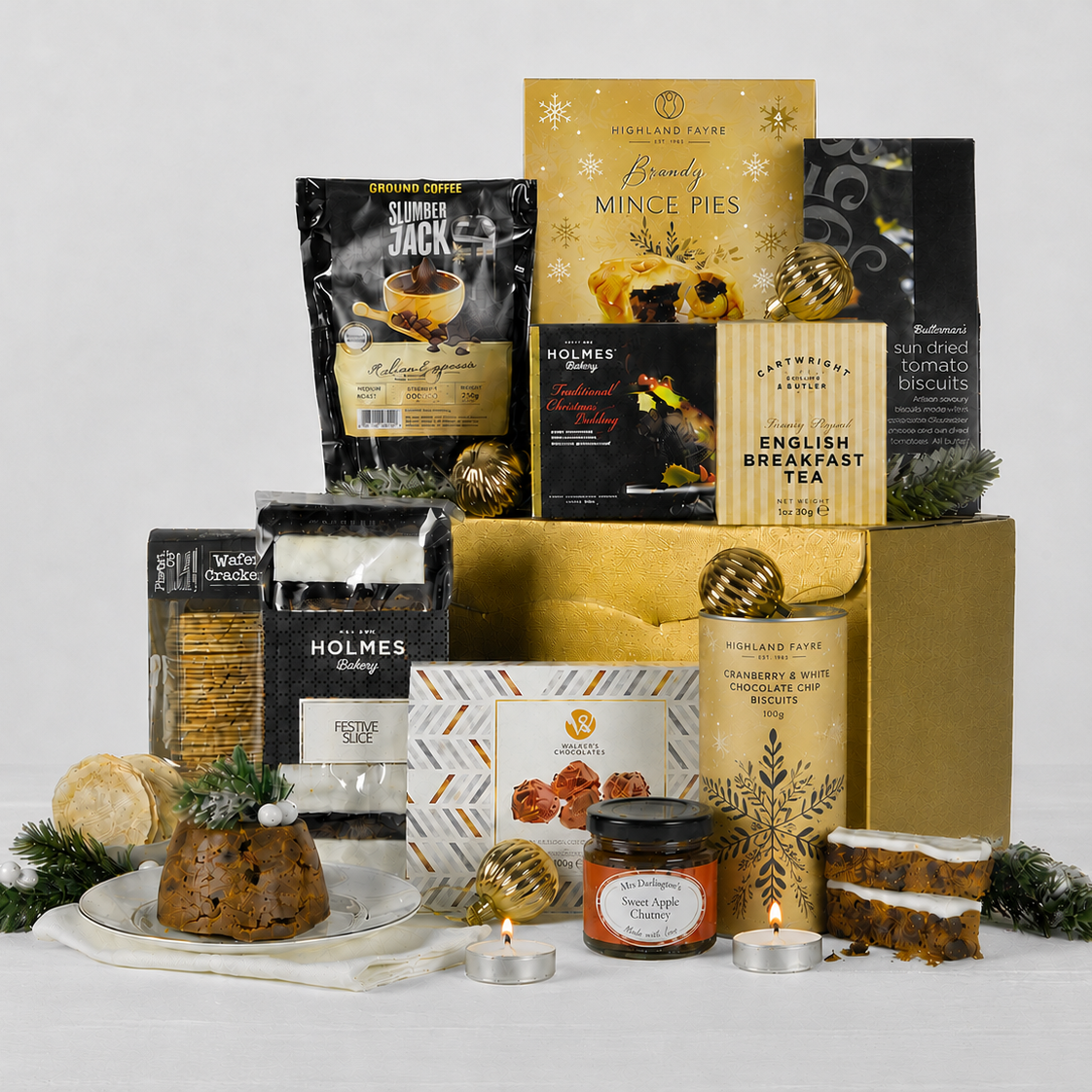 A curated Christmas gift hamper arrangement featuring multiple packaged food and beverage products, including Holmes Bakery, Cartwright & Butler & Carter tea, Slumber Jack, Brandy Mince Pies, styled with greenery and candles in front of a gold gift box on a white background.