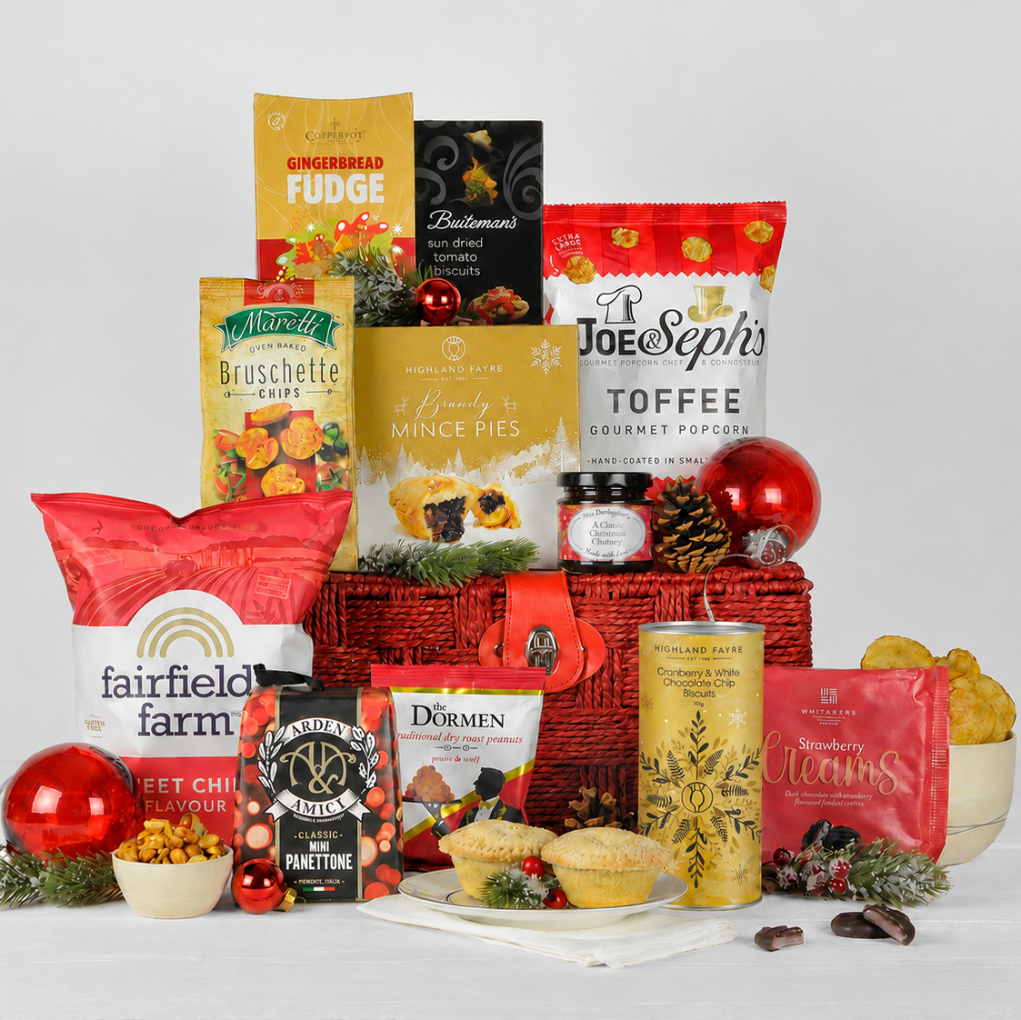 A holiday gift hamper arrangement featuring an assortment of gourmet snacks, sweets, and tea bags styled with red ornaments, pinecones, and mini tarts on a white background.
