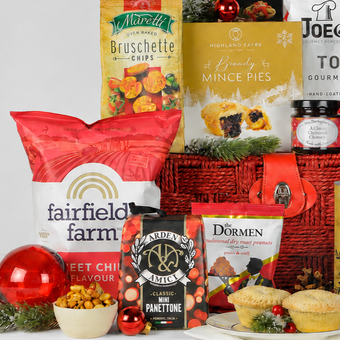 A Christmas-themed gourmet food gift basket filled with assorted treats including a large bag of Fairfield Farm Crisps, a bag of Marretti Bruschette Chips, Highland Faye Brandy Mince Pies, and a jar of Mrs Darlington's Christmas Chutney, arranged with a red wicker basket, red ornaments, and mince pies on a white background.