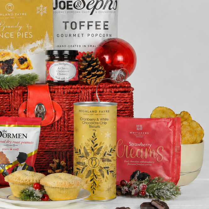 A Christmas-themed food gift basket arrangement on a white surface. The basket is red with a gold clasp and contains a can of Highland Fayre cranberry and white chocolate chip biscuits, a red bag of Whitakers Strawberry Creams, a bag of Joe & Stephs toffee gourmet popcorn.