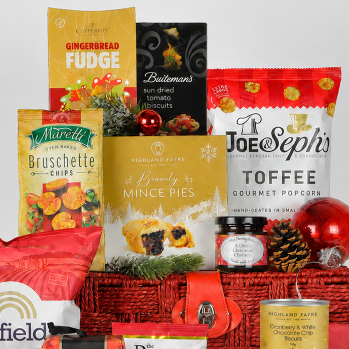 A Christmas-themed gourmet gift basket filled with assorted snacks and treats, including Highland Fayre Brandy Mince Pies, Joe & Seph's Toffee Gourmet Popcorn, and Maretti Bruschette Chips, arranged in a red wicker basket with pinecones and red ornaments.