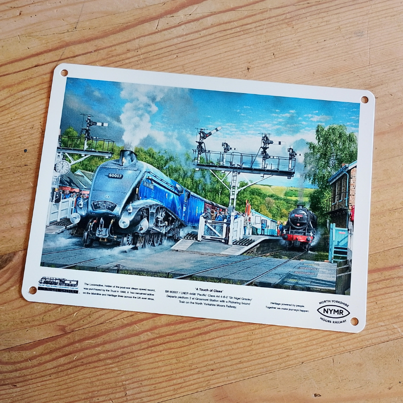 A rectangular printed metal plaque with rounded corners and mounting holes, featuring a colorful illustration of a blue steam train at a station. The plaque is photographed on a light-colored wooden surface.
