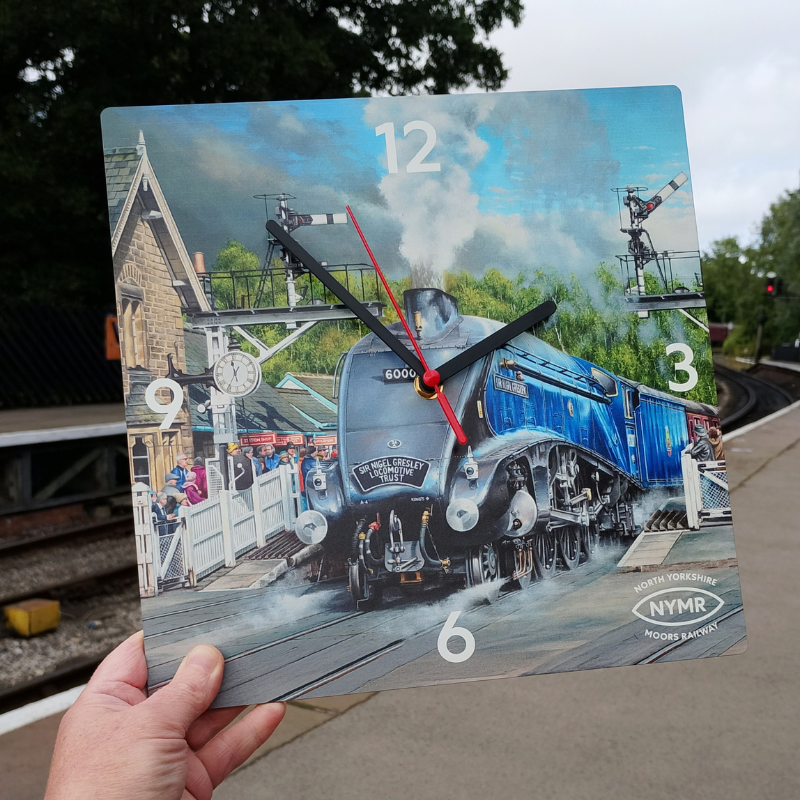 A square wall clock held by a hand, featuring a printed illustration of a blue A4 steam locomotive train at a station. The clock face has white Arabic numerals, black hour and minute hands, and a red second hand.
