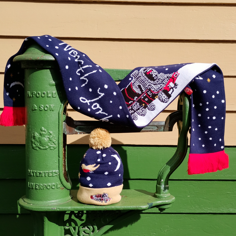 A knitted scarf and bobble hat displayed on station weighing scales. The scarf is navy and white with red detail and fringing. The hat is navy with buff trim and bobble. Trim features embroidered locomotive to match the scarf.