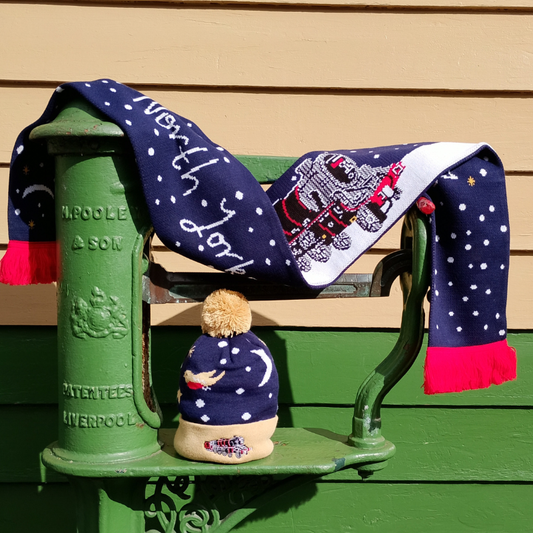 A knitted scarf and bobble hat displayed on station weighing scales. The scarf is navy and white with red detail and fringing. The hat is navy with buff trim and bobble. Trim features embroidered locomotive to match the scarf.