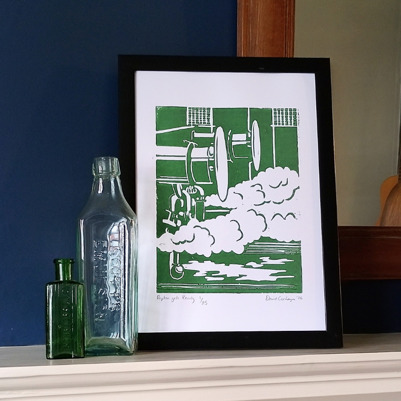 A framed art print featuring a green and white linocut of a steam engine, displayed on a shelf next to two vintage glass bottles against a dark blue wall.