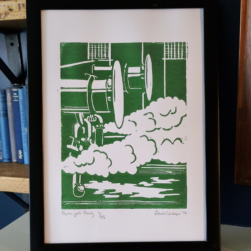 A framed art print featuring a green and white linocut print of a steam engine and smoke, displayed on a shelf next to books.