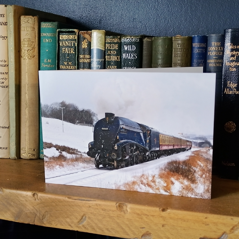 A rectangular greetings card in landscape format, showing the steam locomotive A4 No 60007 Sir Nigel Gresley traveling through a snowy moorland landscape, displayed on a wooden shelf in front of a row of vintage books.