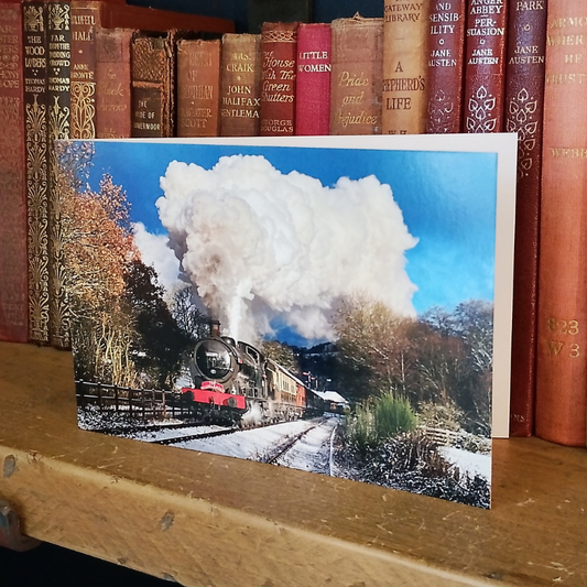 A rectangular photographic greetings card in landscape format showing a steam train traveling along snowy tracks with a large white smoke plume under a blue sky, leaning against a wooden shelf in front of a row of antique books.