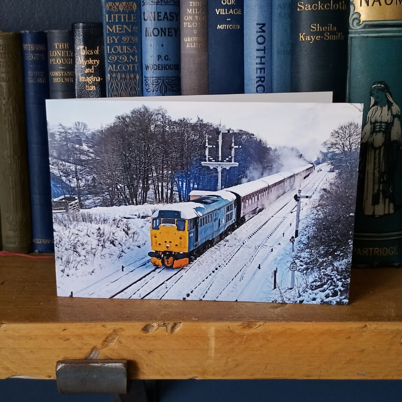 A rectangular greetings card in landscape format showing a snowy winter scene with a diesel engine, leaning on a wooden shelf in front of blue books.