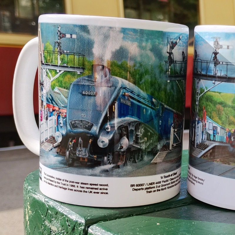 A white ceramic mug featuring a detailed printed illustration of a blue steam locomotive train on a bridge, photographed outdoors on a green painted surface. 