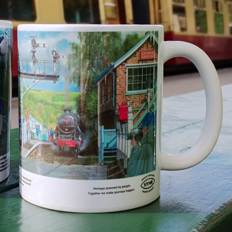 A white ceramic mug featuring a detailed printed illustration of a vintage steam train station scene with a black steam locomotive, brick building, signal equipment, and a person in a green uniform. The mug is photographed on a green tabletop with a blurred train station background.