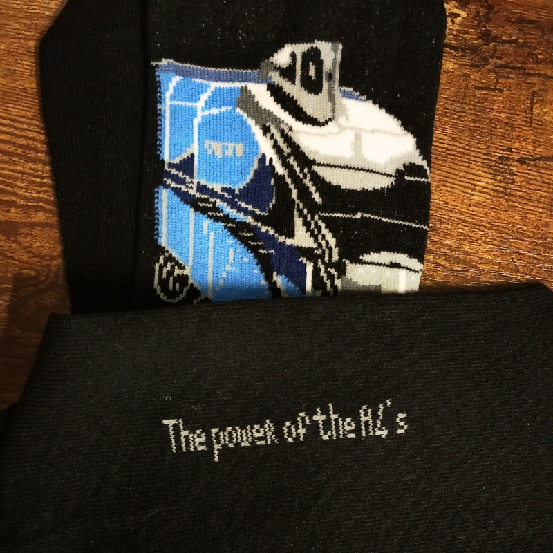 A close-up of a black knit sock with a blue A4 locomotive graphic and the text 'The power of the A4's' embroidered on the bottom, laid flat on a wooden surface.