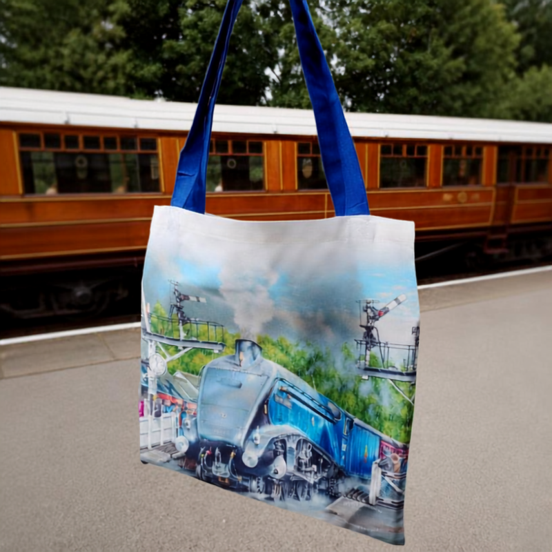 A white canvas tote bag with blue handles, featuring a printed graphic of a blue steam train on a station platform, set against a blurred background of a brown passenger train and green trees.