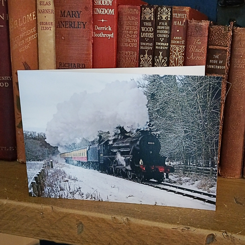 A rectangular greetings card in landscape format showing a snowy winter landscape with a black steam locomotive traveling along tracks, placed on a wooden shelf in front of a row of vintage hardcover books.