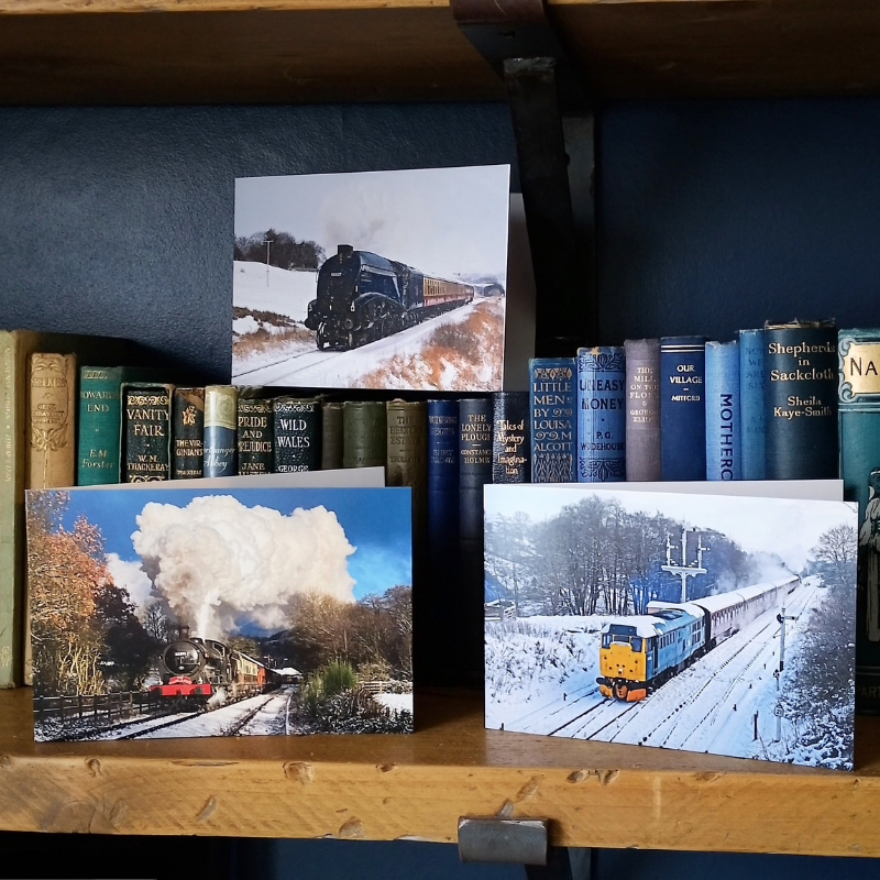 Three greetings cards featuring steam and diesel trains in snowy landscapes, displayed on a wooden shelf next to a row of antique books.