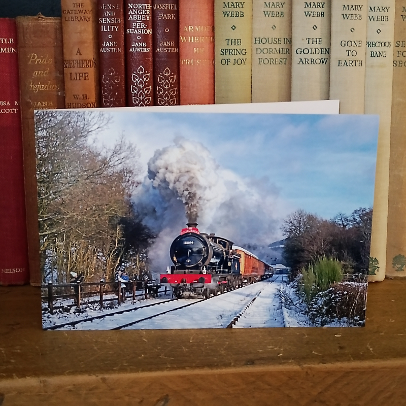A rectangular greetings card in landscape format showing a snowy landscape with a black steam locomotive train emitting smoke, placed on a wooden surface in front of a row of vintage books.