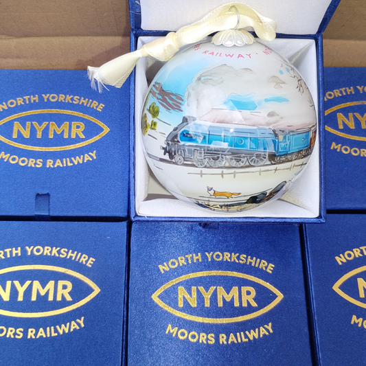 A hand-painted glass Christmas ornament depicting a blue locomotive train on a railway track, presented in its blue gift box with a cream ribbon. The box is surrounded by other blue boxes printed with 'North Yorkshire NYMR Moors Railway'.
