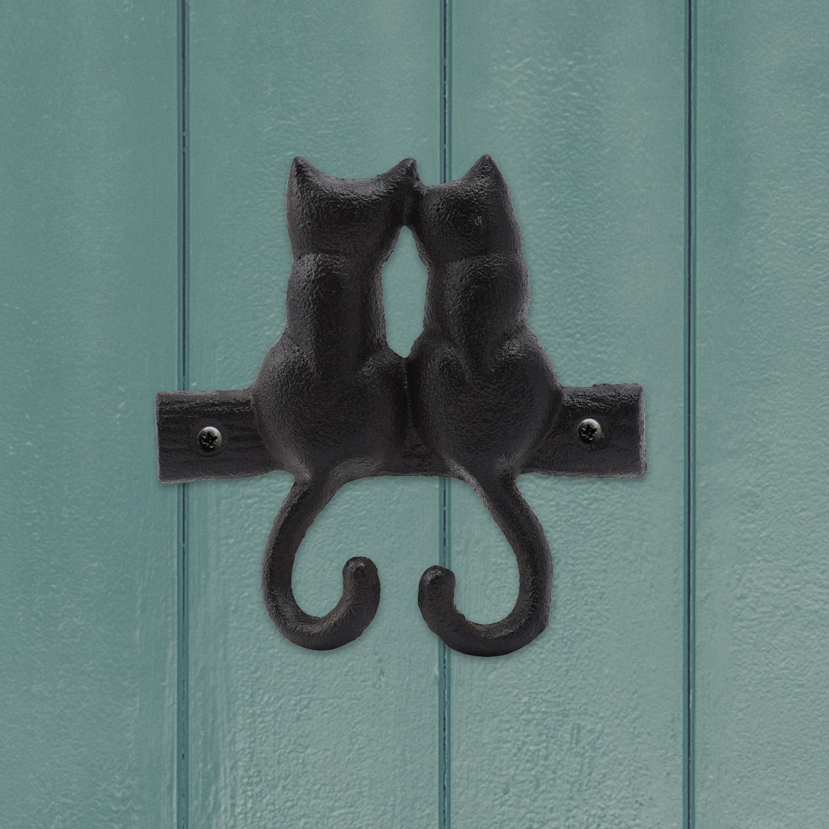 A cast iron hook that is shown screwed to a wooden painted wall. The hooks are made by the curved tails of two cats which are shown from the back.
