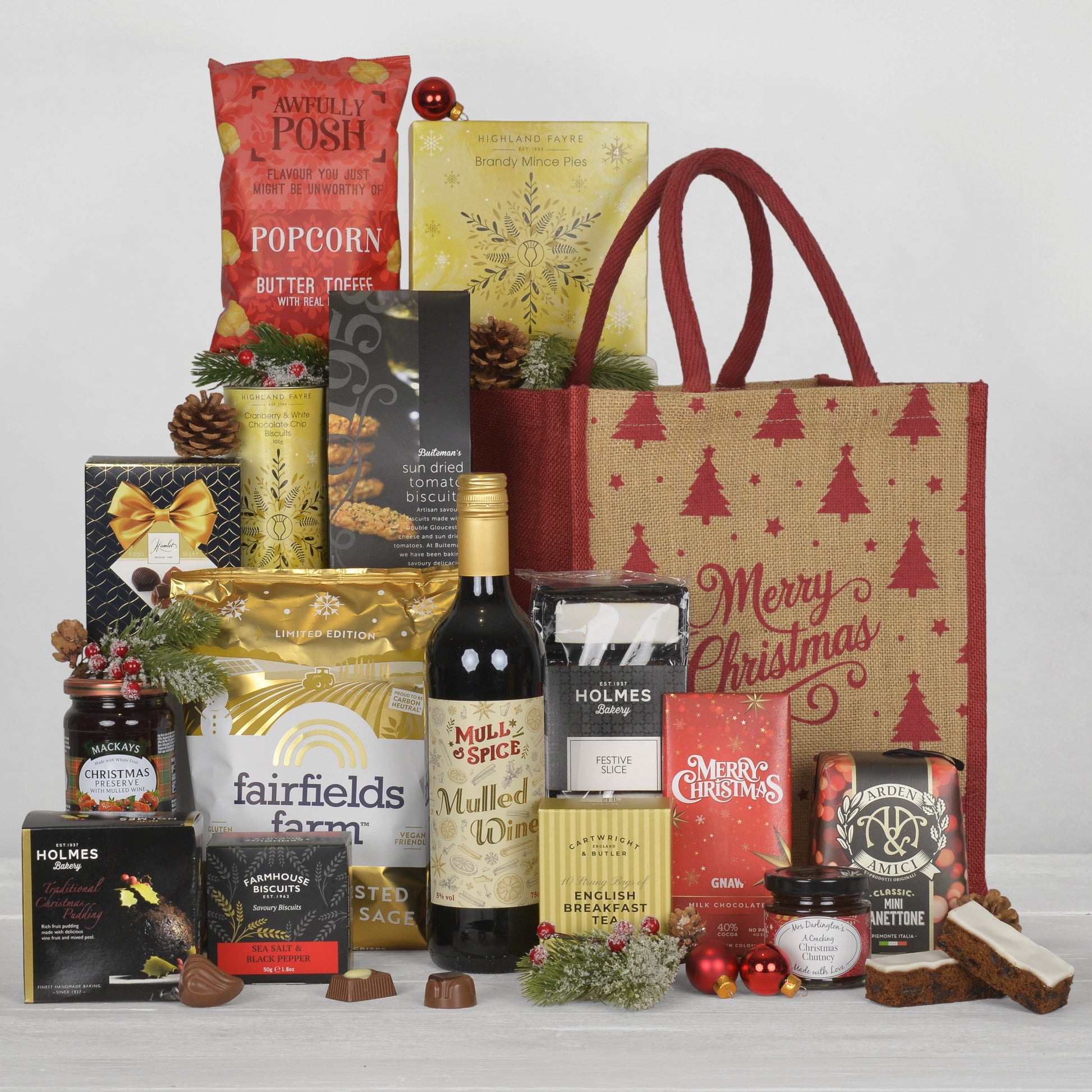 A Christmas gift hamper arrangement featuring a large brown and red jute bag with red Christmas tree prints and 'Merry Christmas' script, filled with assorted packaged treats including popcorn, chocolates, biscuits, jam, and a bottle of Mulled wine.