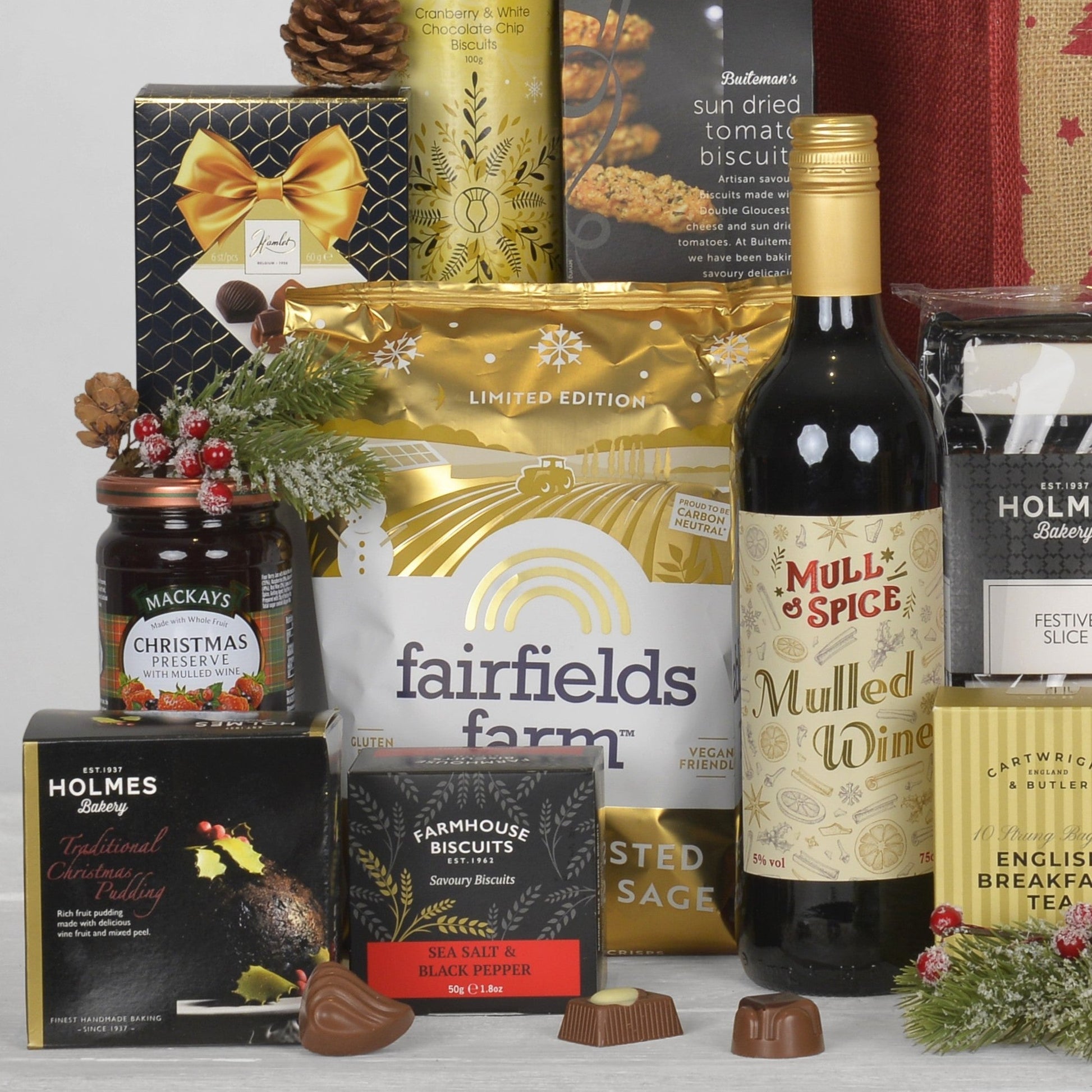 A curated gift hamper arrangement featuring multiple packaged food items: a bottle of Mull & Spice Mulled Wine, a jar of Mackays Christmas Preserve, boxes of Holmes Bakery Christmas Pudding, Farmhouse Biscuits, and other holiday gift boxes and chocolates, styled with pine branches and a gold bow on a white surface.
