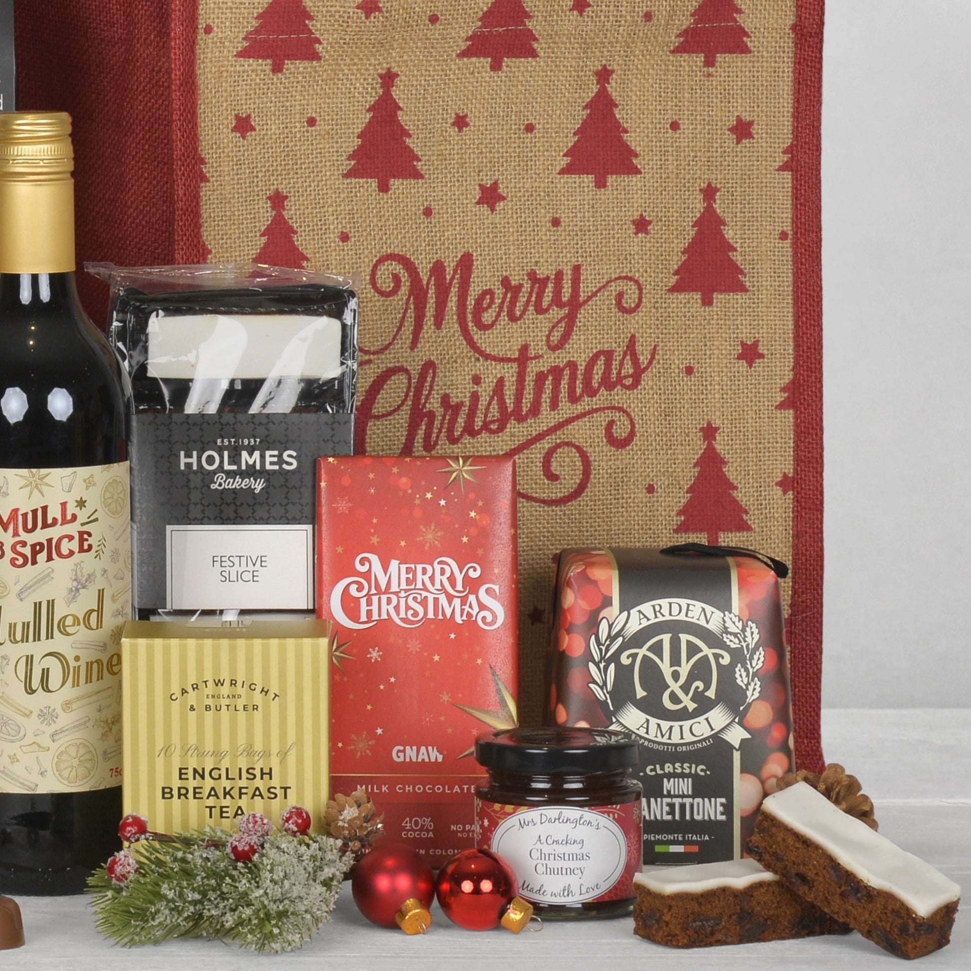 A Christmas-themed gift hamper arrangement featuring a Mull & Spice mulled wine bottle, a box of English Breakfast Tea, a package of Festive Slice, a red Merry Christmas jute bag