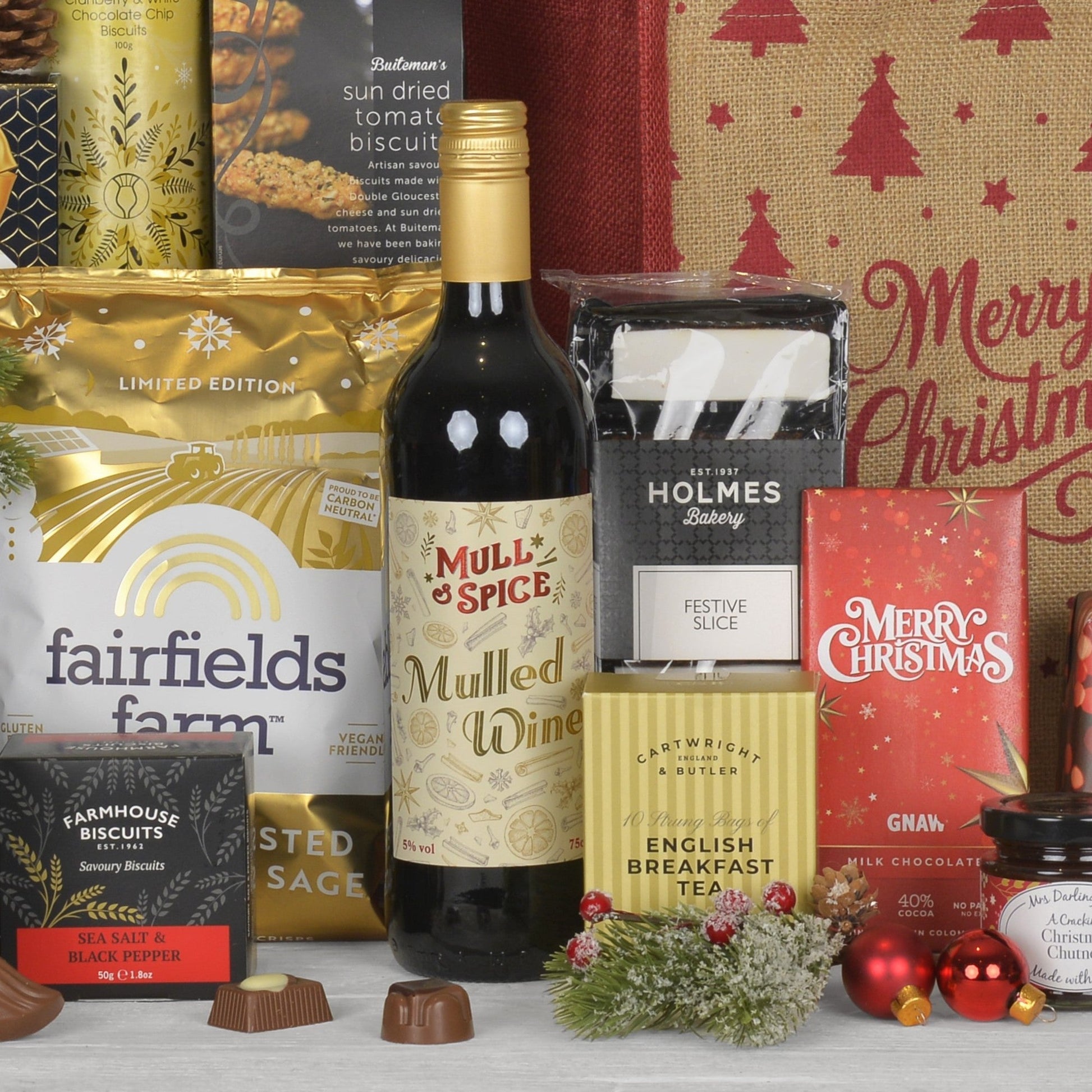 A Christmas hamper gift set featuring a bottle of Mulled Wine, Fairfield Farm crisps, English Breakfast Tea, a box of Holmes Bakery festive slices, Gnaw milk chocolate bar, and other holiday treats arranged on a white surface with a burlap sack and red ornaments in the background.