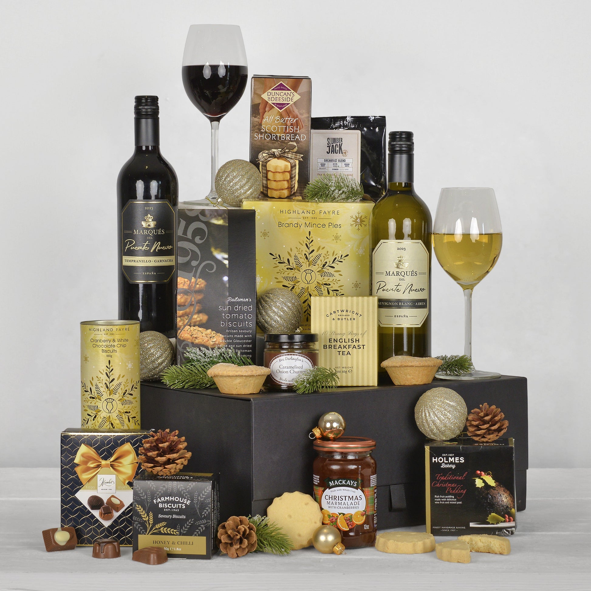 A luxury gourmet gift hamper arranged on a black box, featuring bottles of red and white wine, packaged chocolates, crackers, and small pastries, with two wine glasses and pinecones, photographed on a light gray background.