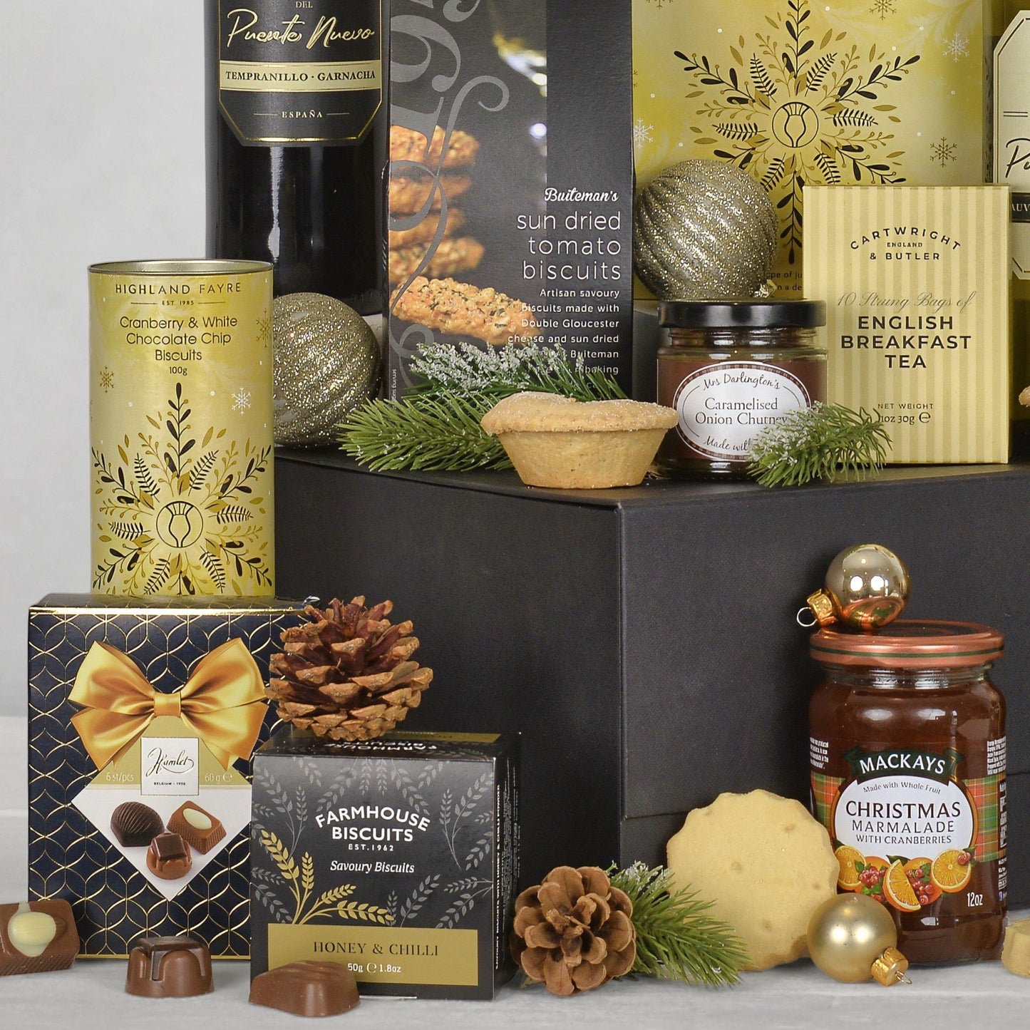 A luxury Christmas gift hamper arrangement featuring an assortment of gourmet treats including Buitman's sun dried tomato biscuits, Highland Fayre Cranberry & White Chocolate Chips, Mackays Christmas Marmalade, and Farmhouse Biscuits, styled with gold ornaments, pinecones, and evergreen branches on a black box.