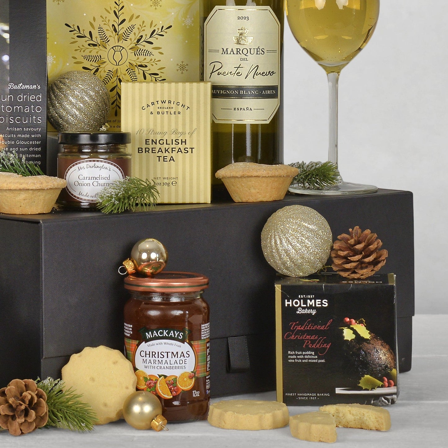 A Christmas gift hamper arrangement featuring a black gift box filled with assorted treats, including a bottle of Marques white wine, a box of English Breakfast Tea, a jar of Mackays Christmas Marmalade, a box of Holmes Christmas Pudding, several cookies, mince pies, and decorative gold ornaments on a light gray background.
