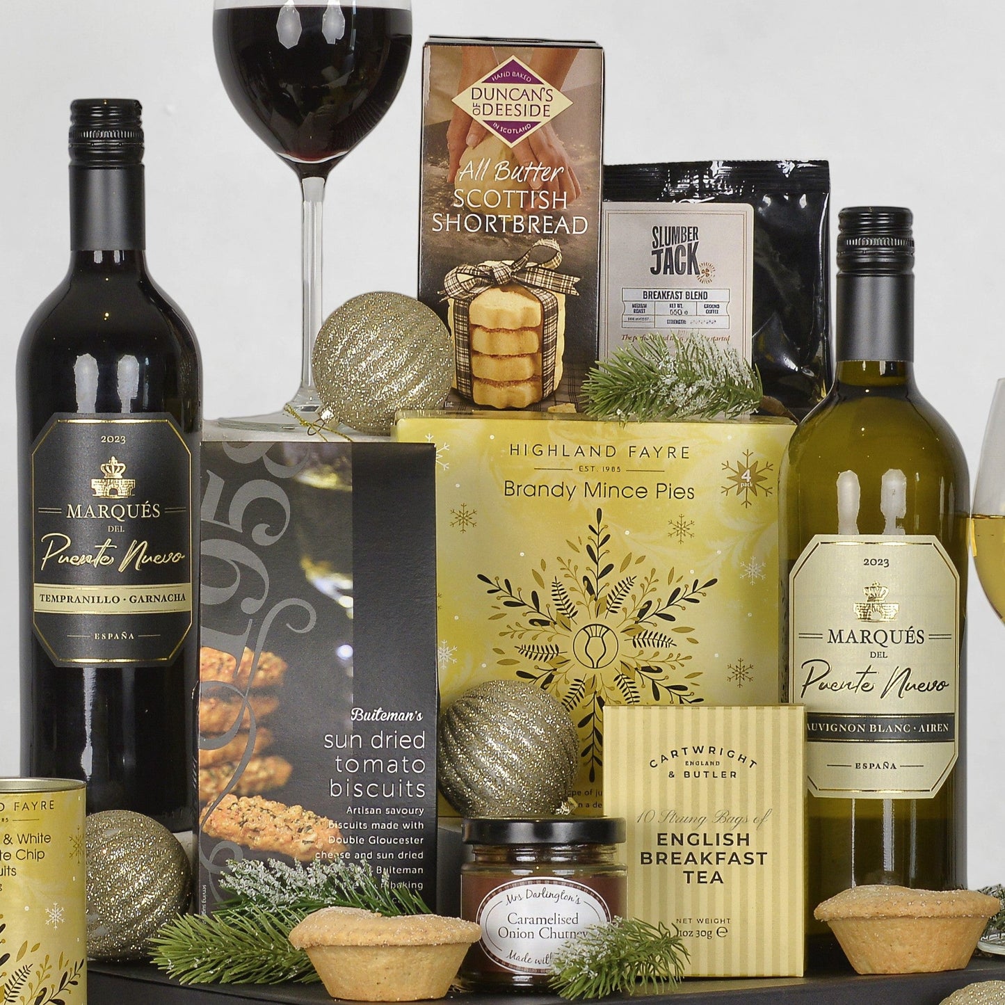 A holiday gift hamper arrangement featuring a bottle of red wine, white wine, Highland Faye Brandy Mince Pies, a bag of Duncan's of Deeside Shortbread, a packet of Slumber Jack Coffee, a box of English Breakfast Tea, styled with gold ornaments, mince pies, and pine branches on a white background.