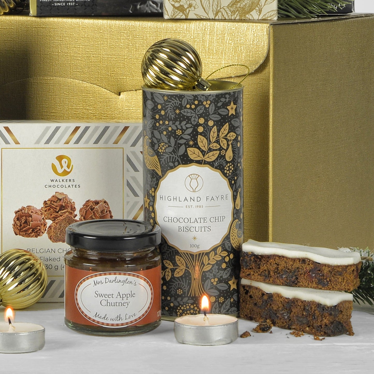 Assorted treats including a jar of sweet apple chutney, Christmas slices, and a box of Highland Fayre chocolate chip biscuits on a table with candles.