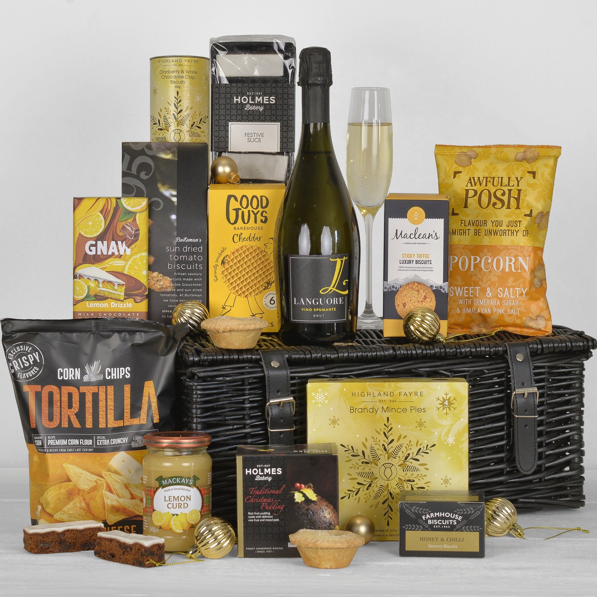 A luxury gourmet hamper gift basket filled with assorted snacks, treats, and a bottle of fizz, arranged in front of a black wicker basket on a light gray background.