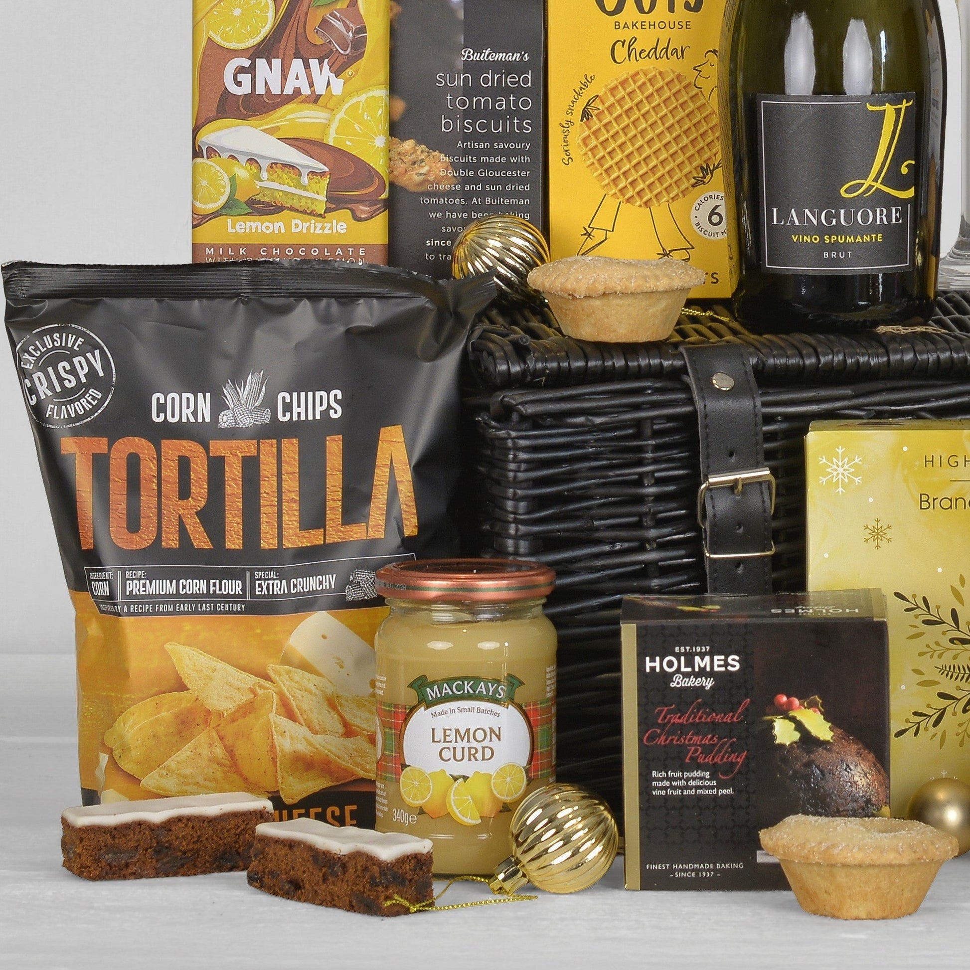 A curated gift basket arrangement featuring a black wicker basket with a large bag of Tortilla Corn Chips, a jar of Mackays Lemon Curd, a bottle of Langeuere Brut wine, a Christmas pudding from Holmes Bakery, and cake and mince pies, styled on a white surface with decorative gold ornaments.