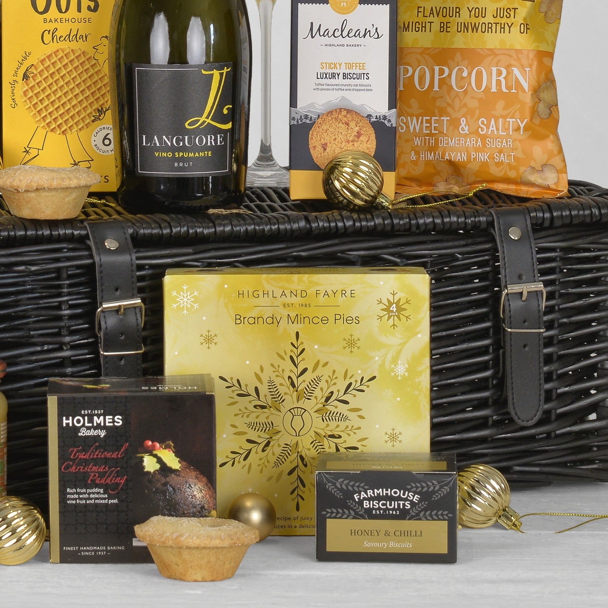 A wicker picnic basket filled with assorted gourmet snacks and treats, including Highland Fayre mince pies, Holmes Christmas Pudding, Maclean's sticky toffee biscuits, Languore wine, and a bag of popcorn, styled with gold ornaments on a white background.