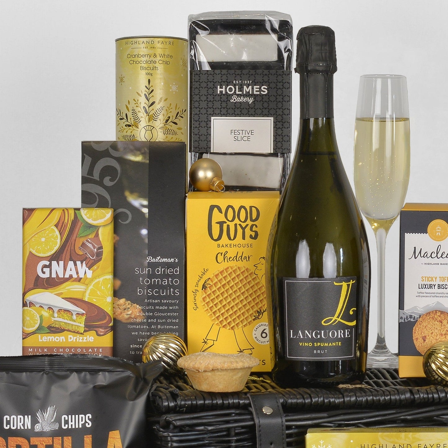 A luxury champagne and chocolate gift basket arrangement featuring a bottle of Langoure Brut, a flute of bubbly, boxed goodies from Good Guys and Holmes, Gnaw lemon drizzle chocolate, and other gourmet treats in a black wicker basket.