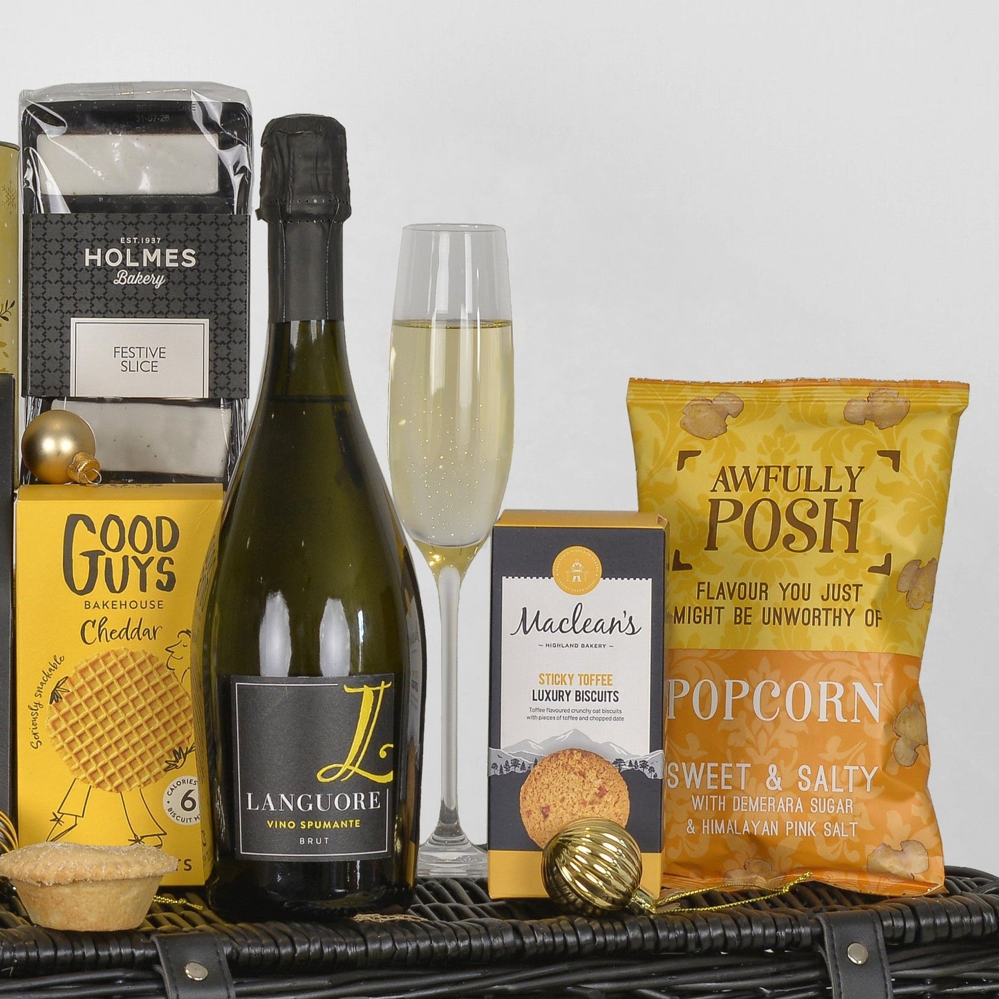 A curated gift basket or hamper arrangement on a black wicker basket, featuring a bottle of Languore Vino Spumante, a flute of bubbly champagne, a bag of Awfully Posh Popcorn, a box of Maclean's Biscuits, a box of Good Guys Cheddar biscuits, and a box of Holmes Bakery Festive Slice.