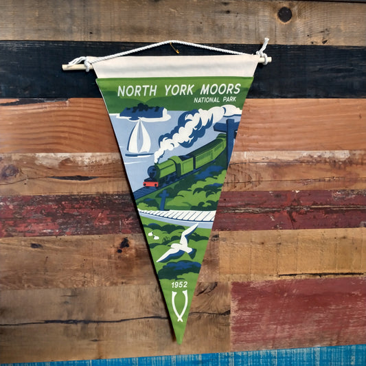 Fabric hanging pennant flag with landscape design and a steam train in the centre. Words across the top say:  NORTH YORK MOORS national park. Predominantly green and blue.