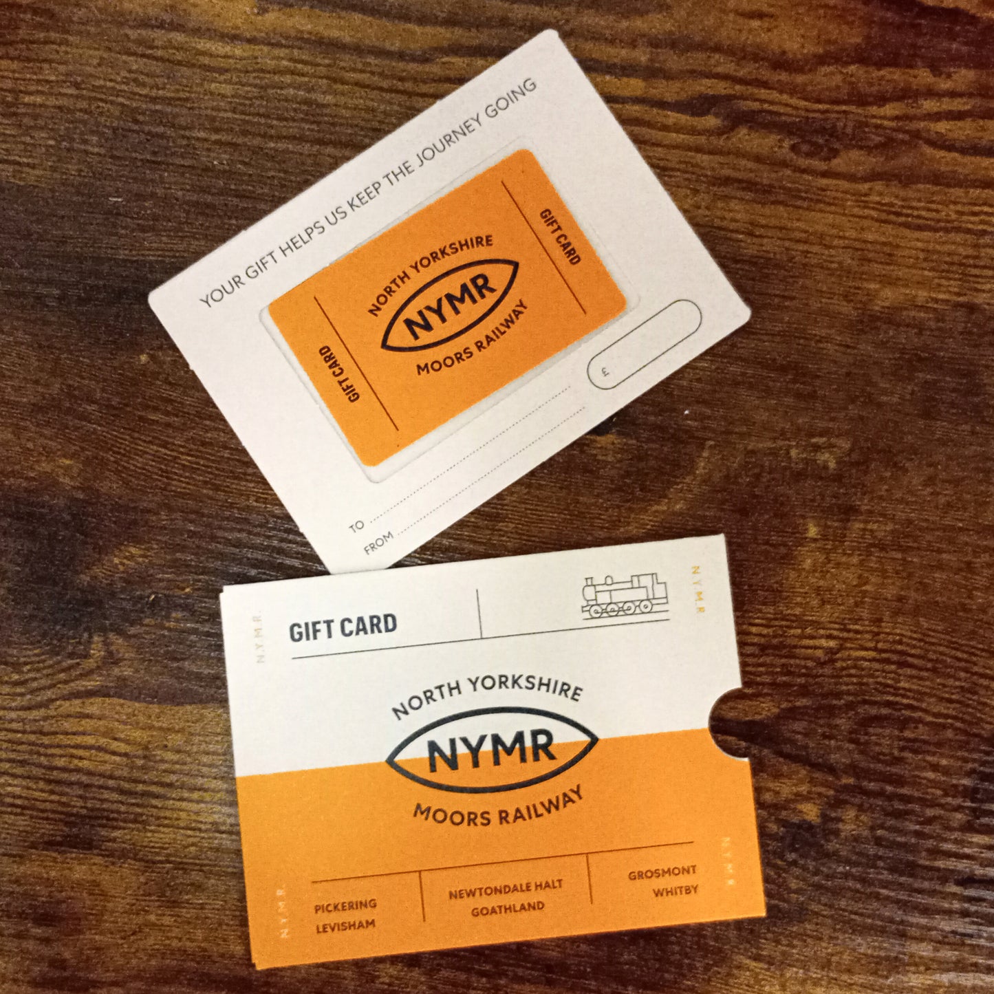 Image shows the credit card style gift card on a card slider which inserts into a card sleeve. Each has the NYMR logo and the words Gift Card on it, plus design details showing an engine and station names.