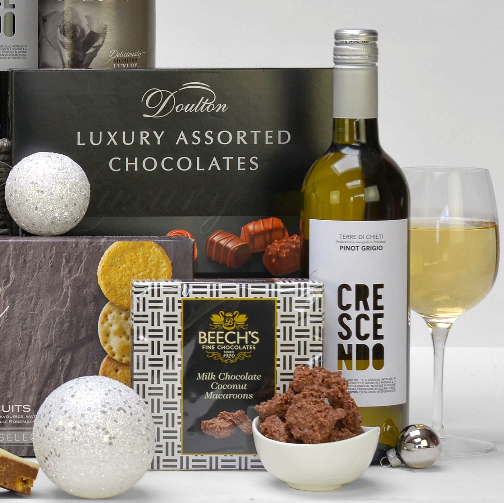 A luxury gift hamper arrangement featuring a bottle of Crescendo Pinot Grigio wine, a box of Doulton luxury assorted chocolates, a box of Beech's Milk Chocolate Coconut Macaroons, and decorative silver ornaments on a white surface.