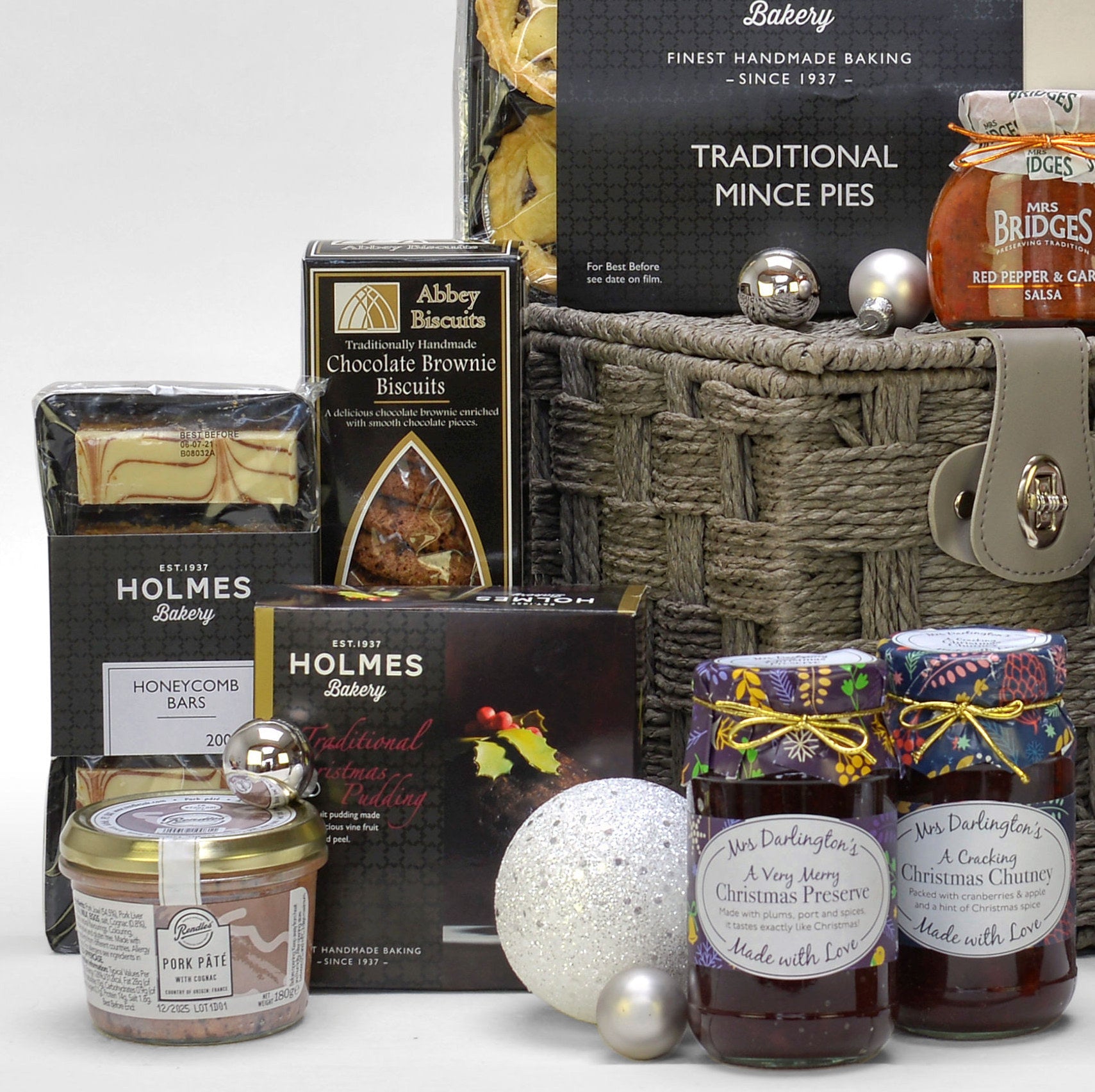 A curated Christmas hamper featuring assorted baking and sweets from brands like Abbey Biscuits, Mrs. Darlington's preserves, Holmes Bakery, arranged around a woven basket with silver ornaments on a white background.