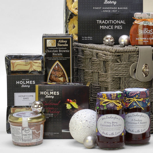 A curated Christmas hamper featuring assorted baking and sweets from brands like Abbey Biscuits, Mrs. Darlington's preserves, Holmes Bakery, arranged around a woven basket with silver ornaments on a white background.