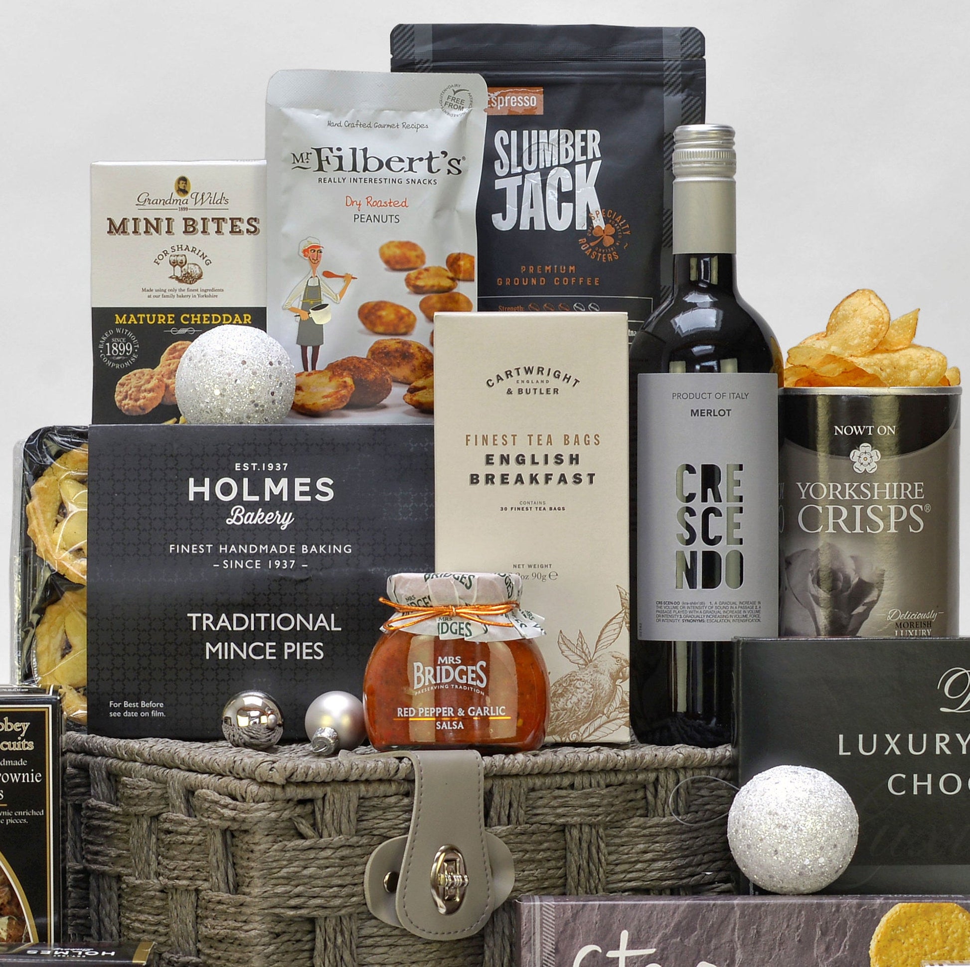 A curated gourmet gift basket arrangement featuring an assortment of food products including a bottle of Merlot wine, a jar of Bridges salsa, a bag of Filbert's peanuts, and various boxed snacks and mince pies, styled on a woven basket with decorative ornaments against a neutral background.