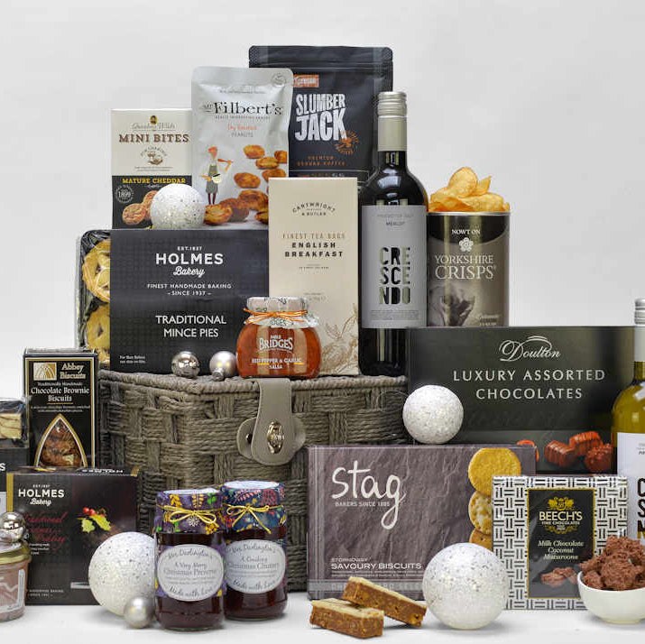 A bottle of red and white wine and Christmas food items such as mince pies, Christmas pudding, Christmas preserve and chutney, tea, coffee, chocolates and savoury snacks displayed with a grey gift basket