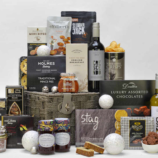 A bottle of red and white wine and Christmas food items such as mince pies, Christmas pudding, Christmas preserve and chutney, tea, coffee, chocolates and savoury snacks displayed with a grey gift basket