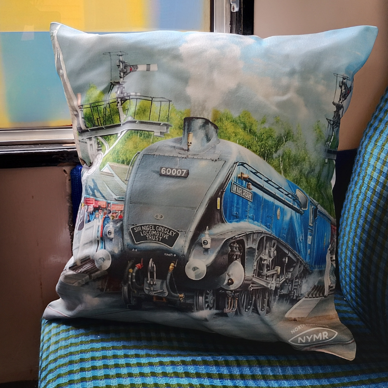 A square decorative cushion featuring a printed illustration of a blue and silver steam locomotive train, resting on a blue and green checkered seat next to a window.